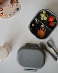 A divided lunchbox with grapes, strawberries, banana slices, olives, a Mushie Trainer Sippy Cup with easy-grip handles, grey cutlery, and a hand holding a silicone palette with colorful circles sit on a white table.
