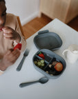 A child sits at a white table, eating a strawberry from a gray lunchbox filled with fruits, next to a fork, spoon, and the Mushie Trainer Sippy Cup with easy-grip handles.