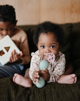 A baby with curly hair uses a FRIGG Baby's First Pacifier Floral Heart (Blush) 4-Pack on the couch, holding a toy, while an older curly-haired child beside them plays with a shape-sorting toy. Both are focused on their activities.