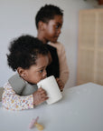 A young child with curly hair drinks from a Mushie Trainer Sippy Cup with easy-grip handles at the table, wearing a floral top and bib. Another child is in the background, slightly out of focus, and a toy rests on the table.