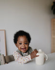 A smiling baby with curly hair sits at a table, holding the Mushie Trainer Sippy Cup, which features easy-grip handles and is non-toxic. The baby wears a floral shirt and bib, with a framed picture and cabinet in the background.