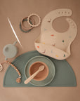 A flat lay of baby feeding items: a green mat, matching plate and bowl with spoon, a beige dinosaur bib, sage green sippy cup, teething rings, and the mushie Silicone Pacifier Clip | Luna—an essential accessory for babies.