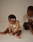 Two young children sit on the floor against a light background. The younger child, in a bow headband and overalls, explores the mushie Flower Press Toy while the older child in stripes looks down, engaging their fine motor skills.