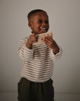 A young child smiles while holding the mushie Phone Press Toy, featuring round buttons for sensory play. The child wears a striped long-sleeve shirt and dark pants, standing against a plain background.