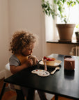 A young child with curly hair sits at a black table, drinking from a cup. On the table are mushie Square Dinnerware Plates with food, a bowl of strawberries, and a fork. Sunlight streams in through a window with a potted plant nearby.