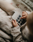 A young child in a light outfit plays with the mushie Phone Press Toy, pressing its round black bubbles for sensory fun, while sitting on a floral-patterned blanket.