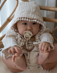A baby in a hooded, textured cream outfit sits on a rattan chair, holding their feet. A FRIGG Daisy Silicone Pacifier from FRIGG is clipped to their clothing, adding comfort amidst soft, neutral-colored decor.