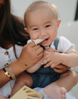 A smiling baby in a white shirt and blue overalls sits on an adult’s lap, chewing on the mushie Finger Toothbrush held by an adult with long brown hair and a gold bracelet.