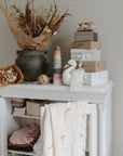 A white cabinet holds folded linens and a mushie Organic Cotton Muslin Swaddle Blanket, topped with gift boxes, a plush goose, colorful stacking cups, and dried flower bouquets against a neutral wall.