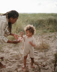 An adult gives a barefoot toddler a pink mushie Snack Cup on a sandy beach with grassy dunes. The toddler, in light casual clothing, examines an object while holding the spill-proof silicone cup.