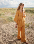 A woman with long blonde hair in a loose mustard-yellow outfit and sandals walks on a sandy beach under cloudy skies, holding a mushie Snack Cup for toddlers and a small beige bag.