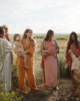 Four women in earth-toned linen relax outdoors, two holding young children engaged with the mushie Phone Press Toy. One woman, visibly pregnant, watches on, creating a serene and natural motherhood vibe.