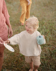 A smiling blond child in a cream sweater and tan shorts holds a blue mushie Snack Cup outdoors on grassy ground, reaching for a plate from an adult. Another person stands in the blurred background.