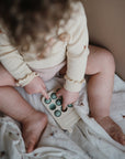 A young child in a cream long-sleeve shirt explores their senses by pressing green bubbles on a mushie Phone Press Toy. The child sits on a patterned blanket, with their face unseen.
