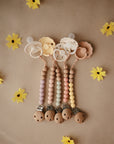 Four mushie Silicone Pacifier Clips | Eva in pastel colors, with food-grade silicone beads and clips, are lined up on brown paper and surrounded by yellow artificial flowers.
