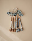 Four mushie "Silicone Pacifier Clip | Eva" in neutral and blue tones with beaded silicone and wooden accents are displayed in a row on a beige background, highlighting stylish and safe pacifier accessories.
