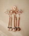 Four mushie Silicone Pacifier Clips | Luna are displayed on a neutral background. Each features food-grade silicone and wooden beads in soft pastel tones, blending function with style for the perfect baby accessory.