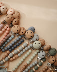 The mushie Silicone Pacifier Clip | Hera features food-grade silicone and wooden beads in soft, neutral colors, arranged neatly on a beige surface for a clean, minimalist look.