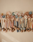 A row of mushie Silicone Pacifier Clip | Eva, featuring food-grade silicone and wooden beads in pastel colors, hangs on a light beige wall as stylish and safe pacifier accessories.