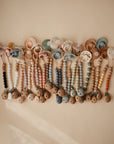 The mushie Silicone Pacifier Clip | Hera features colorful beaded designs and wooden clasps, crafted from food-grade silicone, and arranged on a beige background for a charming, minimalist look.