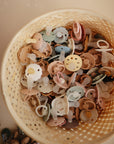 A top view of a basket filled with FRIGG Moon Silicone Pacifier 2-Pack sets in pastel colors, featuring medical grade silicone and adorable flower or smiley designs—essential baby items from FRIGG.