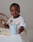 A young child in a white outfit sits at a white table holding a blue sippy cup and a FRIGG Daisy Natural Rubber Pacifier, with a colorful palette-shaped toy nearby. A wicker-back chair is partly visible in the background.