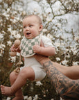A smiling baby wears a checkered bib and the mushie Silicone Pacifier Clip | Hera, held up by tattooed adult hands among blooming white flowers on tree branches.