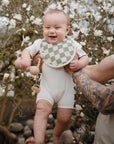A smiling baby in a white romper and checkered bib, secured with the mushie Silicone Pacifier Clip | Hera, is held by a tattooed adult in front of a flowering tree.