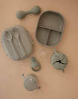 A set of sage green silicone baby feeding items on a tan background, featuring Mushie’s Trainer Sippy Cup with easy-grip handles, a divided plate, snack cup, spoon and fork set, teether, and a pacifier with cover.