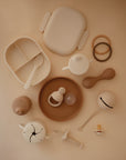 A flat lay of beige and brown baby items, including plates, spoons, a pacifier, teething rings, a bib, a rattle, and the Mushie Trainer Sippy Cup with easy-grip handles, arranged neatly on a neutral background.