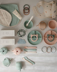 A flat lay of pastel baby feeding items, including mushie’s Organic Cotton Muslin Burp Cloth 2-Pack, with bibs, plates, bowls, cups, spoons, teething rings, snack containers, cream tubes—all neatly arranged on a light surface.