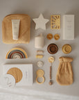 A selection of neutral baby essentials, including Mushie’s Organic Cotton Bath Mitt, sensitive skin lotion, towel, star toy, pacifier, stacking rings, teether, and rattle arranged on a beige background.