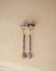 Two mushie Silicone Pacifier Clips | Eva hang side by side on a beige background; one features light blue food-grade silicone beads, while the other has a gray clip with mixed wooden and gray pacifier accessories.