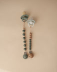 Two mushie Silicone Pacifier Clips | Eva, made from food-grade silicone, are shown on a beige background. One has green and cream beads, the other grey and wooden beads, offering safe and stylish options for baby accessories.