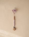A mushie Silicone Pacifier Clip | Eva with alternating purple and wooden beads secures a purple pacifier, displayed on a beige background.