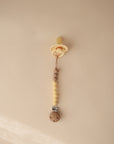 The mushie Silicone Pacifier Clip | Eva, featuring a beige beaded design with a round wooden clip, hangs on a light tan wall—an elegant addition to your pacifier accessories collection.