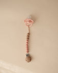 The mushie Silicone Pacifier Clip | Eva, featuring wooden and food-grade silicone beads, holds a pink baby pacifier and is displayed on a light beige surface.
