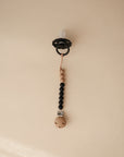 The mushie Silicone Pacifier Clip | Eva features a black pacifier attached to a food-grade silicone clip with alternating black and wooden beads, shown against a plain beige background.