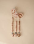 Three pink mushie Eva Silicone Pacifier Clips with beaded food-grade silicone and wood details are arranged side by side on a beige background, offering stylish and practical pacifier accessories.