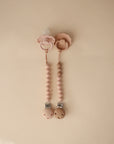 Two mushie Silicone Pacifier Clips | Eva, one with food-grade silicone beads and the other with wooden beads, are placed side by side on a beige background.