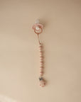 The mushie Silicone Pacifier Clip | Luna, featuring a pink pacifier and beaded food-grade silicone clip, is displayed vertically on a plain light beige background.