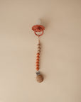 The mushie Silicone Pacifier Clip | Eva securely holds a red baby pacifier with alternating wooden and orange food-grade silicone beads, displayed on a neutral beige background.