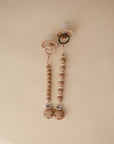 Two FRIGG Andersen Fairytale Silicone Pacifiers—one scalloped, one plain—with round beaded wooden clips and metal clasps are displayed on a light beige background. Made in Denmark by FRIGG.