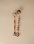 Two mushie Silicone Pacifier Clip | Luna clips, made with round wooden beads and metal clasps, are attached to food-grade silicone pacifiers and photographed on a beige background.