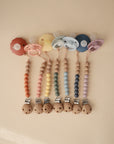 Six mushie Silicone Pacifier Clips | Eva, featuring food-grade silicone beads and wooden holders, are arranged in a row on a beige background, each topped with a pastel pacifier.