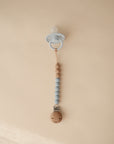The mushie Silicone Pacifier Clip | Eva features wooden and blue food-grade silicone beads with a light blue pacifier, shown on a beige background.