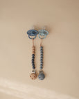 Two mushie Silicone Pacifier Clip | Hera sets, each with a blue pacifier, hang side by side on a beige background. The minimalist clips feature round wooden ends and food-grade silicone beads in blue and natural wood tones.