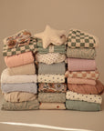 Three neat stacks of pastel blankets and quilts, topped with a star-shaped pillow, are arranged beside cozy mushie Organic Cotton Muslin Burp Cloths 2-Pack, all displayed against a beige background.