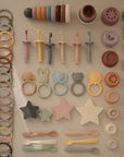 Mushie Finger Toothbrush, along with colorful animal- and star-shaped silicone baby toys, stacking cups, bibs, spoons, pacifiers, and teethers, are arranged neatly in rows on a beige background.