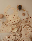 A flat lay of baby feeding items, including the mushie Silicone Pacifier Clip | Luna, a fox-print bib and placemat, white bowl with wooden spoon, tan sippy cup, pacifier, and matching muslin blanket—perfect baby accessories made from food-grade silicone.